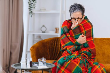 Kendini hasta hisseden bir kadın, sıcak bir battaniyeye sarılmış, evde dinlenirken üşütüp kokluyor.