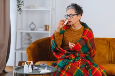 Üşütmüş ya da grip olmuş bir kadın evde sıcak bir battaniyeye sarılı iken burun spreyi kullanıyor.