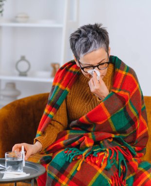 Kadın kendini kötü hissediyor, kanepeye oturmuş battaniyeye sarılmış ve burnunu mendille siliyor.