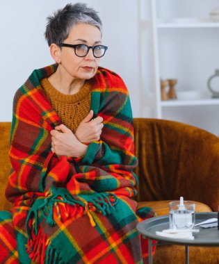 Üst düzey kadın battaniyeye sarılı olarak koltukta titriyor. Soğuk algınlığı ya da grip belirtileri yüzünden evde dinleniyor ve iyileşiyor.