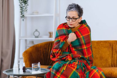 Yaşlı kadın üşümüş, sıcak bir battaniyeye sarılmış, evinde ateşle ya da griple boğuşuyor.
