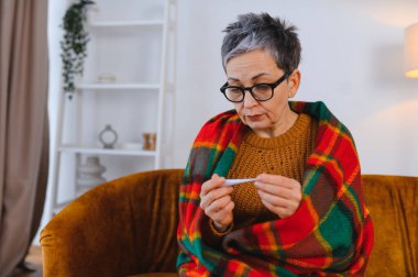 Sıcak bir battaniyeye sarılmış, kanepeye oturmuş, termometreye bakıyor, soğuk algınlığı ve grip belirtileri gösteriyor.