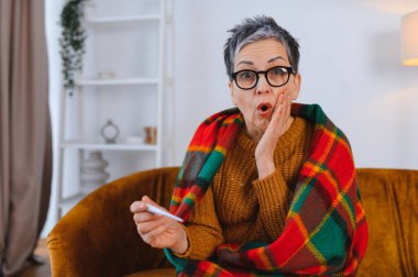 Yaşlı kadın grip belirtilerinden rahatsız, battaniyeye sarılmış, termometreye bakıyor.