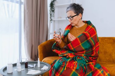 Yaşlı kadın kendini hasta hissediyor, battaniyeye sarılmış, suyla birlikte bir hap alıyor, çeşitli soğuk algınlığı ve grip ilaçlarıyla sarılmış.