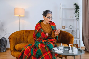 Yaşlı kadın battaniyeye sarılıp ateşini ölçüyor. Soğuk algınlığı, grip ve hastalık belirtileri gösteriyor.