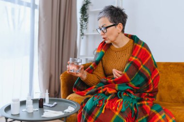 Sıcak battaniyeye sarılmış, soğuk algınlığı geçiren, elinde bir bardak su tutan ve ilaç yazan olgun bir kadın.