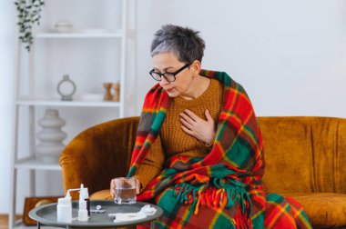 Kanepeye sarılmış, elinde bir bardak su tutan ve ilaç alan olgun bir kadın. Hastalık belirtileri yüzünden göğüs ağrısı çekiyor.