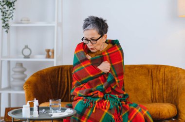 Yaşlı kadın kendini iyi hissetmiyor, sıcak bir battaniyeye sarılmış ve evdeki kanepede titriyor, masada grip ilacıyla.