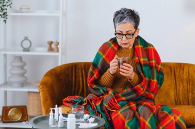 Yaşlı kadın soğuk semptomlarla savaşıyor, sıcak bir battaniyeye sarılmış, yakınlardaki masada ilaçlarla çay içiyor.