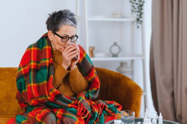 Kareli battaniyeye sarılı yaşlı bir kadın evde sıcak çay içiyor, mevsimsel soğuk ve hastalıkla baş ediyor.