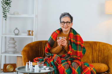 Battaniyeye sarılmış, gülümseyen ve sıcak bir içecek tutan olgun bir kadın. İlaçlar yakınlardaki bir masada.