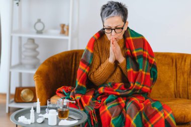 Yaşlı bir kadın, ilaç ve mendillerle sarılı bir battaniyeye sarılmış, titriyor ve öksürüyor.