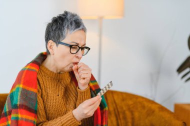 Soğuk algınlığı veya grip geçiren yaşlı bir kadın, sıcak bir battaniyeye sarılmış, öksürürken ilaç tutuyor.