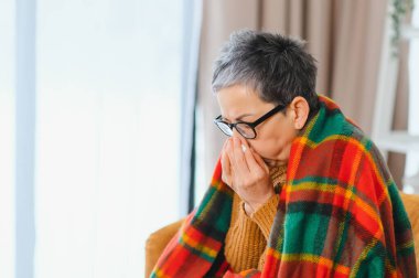 Üst düzey bir kadın sıcak bir battaniyeye sarılı halde burnunu mendille kapatıyor. Soğuk algınlığı ya da evde grip belirtileri gösteriyor.