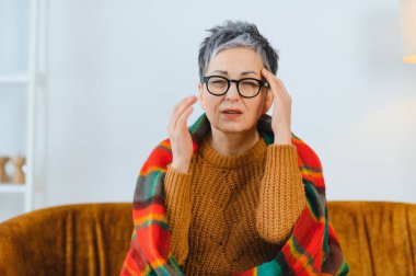 Yaşlı kadın şiddetli migren ağrısı çekiyor, tapınaklara dokunuyor, sıcak bir battaniyeye sarılı kendini iyi hissetmiyor.