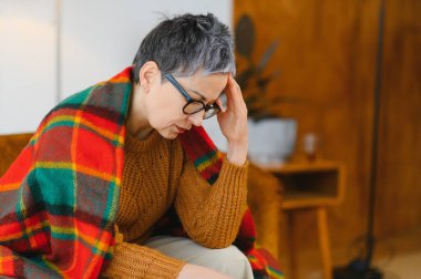 Yaşlı gri saçlı kadın battaniyeye sarılıp dinlenirken kronik baş ağrısı, stres ya da diğer hastalıklar yaşıyor.