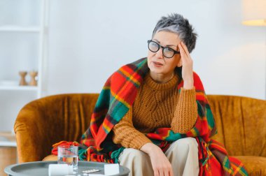 Yaşlı bir kadın baş ağrısı ya da migren yüzünden acı çekiyor. Yanında ilaçlarla kanepeye sarılmış bir şekilde oturuyor.