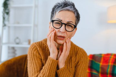 Şiddetli diş ağrısı çeken yaşlı bir kadın, yanağına dokunuyor. Diş sorunları ve oral sağlık sorunları kavramı