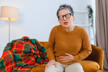 Kanepede oturan yaşlı bir kadın, karın ağrısı çekiyor. Elleri ağrıyan karnına bastırıyor.