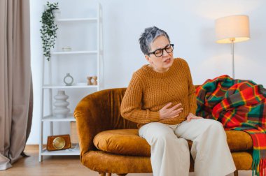 Oturma odasındaki kanepede otururken karın ağrısı, kramp ve hazımsızlık çeken yaşlı bir kadın.