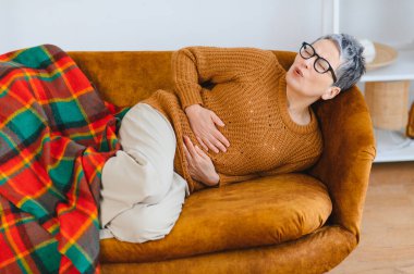 Olgun bir kadın kahverengi bir kanepede uzanıyor, elleriyle karnını dinlendiriyor, karın ağrısı, rahatsızlık ya da hazımsızlık çekiyor.