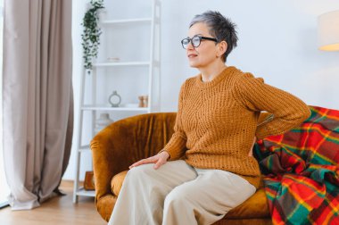 Yaşlı kadın sırt ağrısı çekiyor, belini tutuyor ve kanepede otururken kendini rahatsız hissediyor.