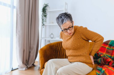 Yaşlı bir kadın sırt ağrısı çekiyor, bir koltukta oturuyor, kederli bir ifadeyle yan tarafını tutuyor.