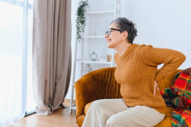 Yaşlı kadın sırt ağrısı çekiyor, bel bölgesini tutuyor, evdeki koltukta oturuyor, rahatsızlık ve gerginlik hissediyor.
