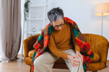 Yaşlı bir kadın kanepede oturuyor, rahatsız hissediyor ve eklem sorunları yüzünden sıcak bir battaniyeye sarılmış, ağrıyan dizini tutuyor.