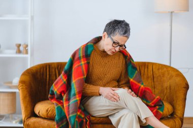 Kanepede otururken diz ağrısı çeken yaşlı bir kadın eklem iltihabı semptomları, yaşlılık hastalığı ve rahatsızlık yaşıyor.