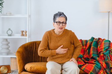 Kanepede oturan bir kadın, göğsünü rahatsızlık içinde tutuyor, kalp ağrısı, endişe ve sağlık sorunları belirtileri gösteriyor.