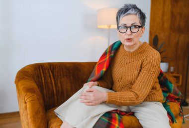 Ağır diz ağrısı çeken son sınıf öğrencisi bir kadın, rahat bir evde koltukta oturuyor, ağrıyan dizine dokunuyor.