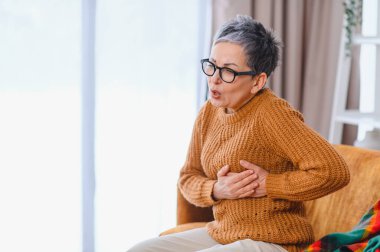 Ağır göğüs ağrısı çeken yaşlı bir kadın, kalp bölgesini sıkıntılı bir ifadeyle tutuyor, muhtemelen kalp krizi geçiriyor.