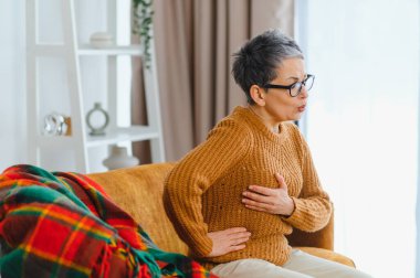 Ağır göğüs ve yan ağrıları olan yaşlı bir kadın, iyi görünmüyor ve evdeki bir kanepede nefes nefese.
