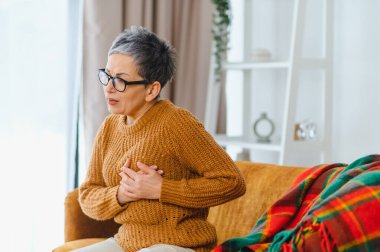 Kanepede oturan olgun bir kadın, göğsünü tutuyor, şiddetli bir acı hissediyor. Kalp krizi ya da anjin belirtileri gösteriyor.