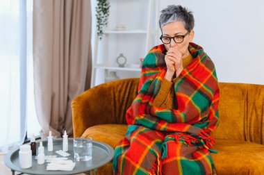 Kadın kendini iyi hissetmiyor. Soğuk algınlığı ve grip belirtileri gösterirken battaniyenin altında titriyor. İlaçlar yakınlardaki bir masada.