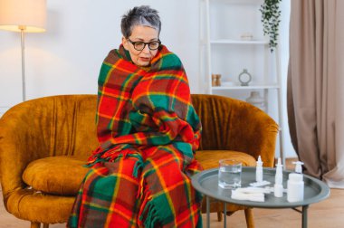 Yaşlı kadın kendini iyi hissetmiyor, sıcak bir battaniyenin altında titriyor. İlaçlar, su ve mendiller yakınlardaki bir masada.