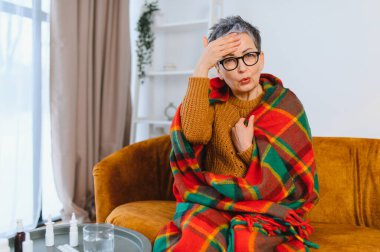 Olgun bir kadın kendini kötü hissediyor ve hastalıktan muzdarip, evinde baş ağrısı ve ateşi var, battaniyeye sarılmış.