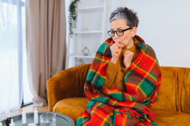 Yaşlı kadın soğuk algınlığı ya da grip yüzünden kendini iyi hissetmiyor. Kanepeye sarılmış, masada ilaç var.