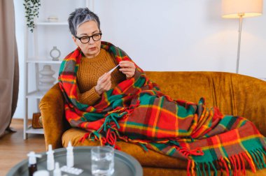 Üst düzey bir kadın kanepeye sarılıp ateşini ölçüyor. Hastalık ve ateşle boğuşuyor.