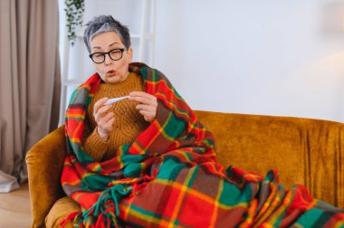 Kıdemli bir kadın, koltuğunda sıcak bir battaniyeye sarılı dijital termometreyi kontrol ederken titriyor ve endişeli görünüyor.