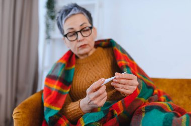 Üst düzey bir kadın sıcak bir battaniyeyle kendini örtüyor, kanepeye oturuyor ve vücut ısısını termometreyle ölçüyor.