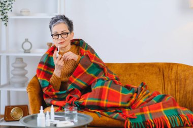 Yaşlı kadın soğuk algınlığı semptomlarından rahatsız. Sıcak bir battaniyenin altında kanepede dinlenirken burun spreyi kullanıyor.