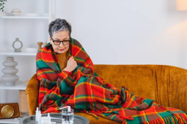 Yaşlı kadın titriyor ve battaniyeye sarılıyor, soğuk algınlığı, grip veya mevsimsel hastalıktan kurtulmak için evde kalıyor.