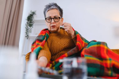 Yaşlı kadın kendini iyi hissetmiyor, evde battaniyeye sarılmış, ilaç alıyor ve hastalık belirtilerini kontrol ediyor.