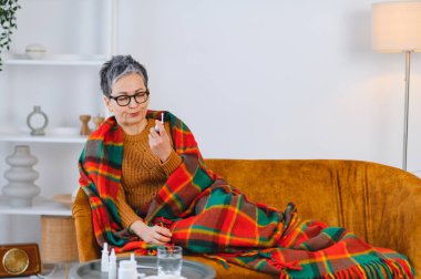 Kanepede oturan yaşlı bir kadın, battaniyeyle örtülmüş, nezle ya da alerji ilacı olarak burun spreyi kullanıyor.