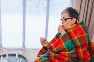 Yaşlı kadın evde battaniyeye sarılmış, grip ya da soğuk algınlığı için ilaç alıyor, hastalık sırasında semptomları ve kendine bakımı gösteriyor.