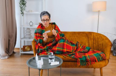 Yaşlı kadın kendini hasta hissediyor. Kanepede battaniyeye sarılı grip belirtileri var. Elinde bir paket ilaç var.