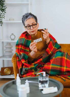 Yaşlı kadın kendini iyi hissetmiyor, battaniyeye sarılmış, soğuk algınlığını veya grip belirtilerini tedavi etmek için ilaç alıyor.