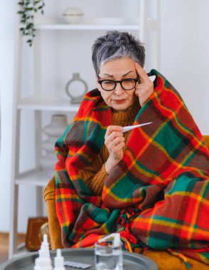 Evde elinde termometreyle oturan yaşlı bir kadın soğuk algınlığı ve ateş belirtileri göstererek kendini hasta hissediyor.